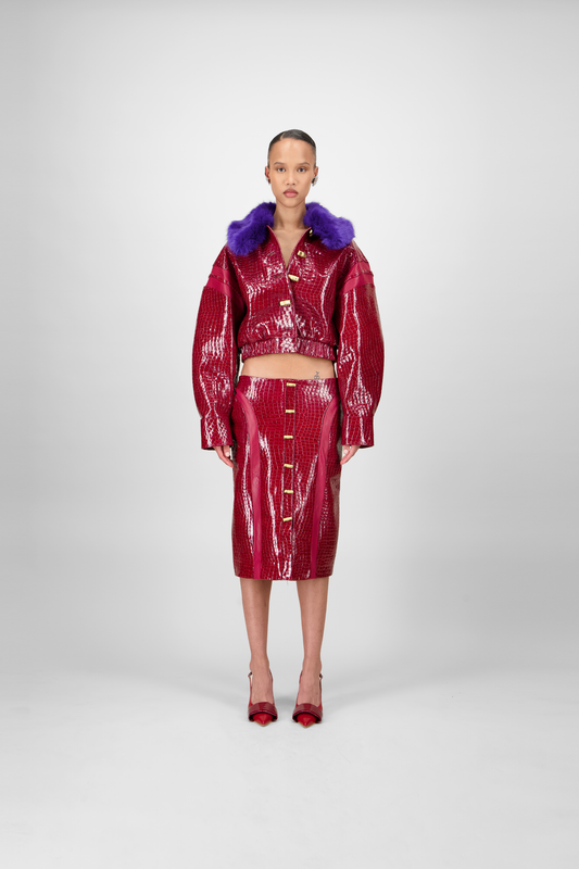 A woman model wearing a red crocodile bomber jacket with a detachable purple faux fur collar and metallic bamboo-like buttons.