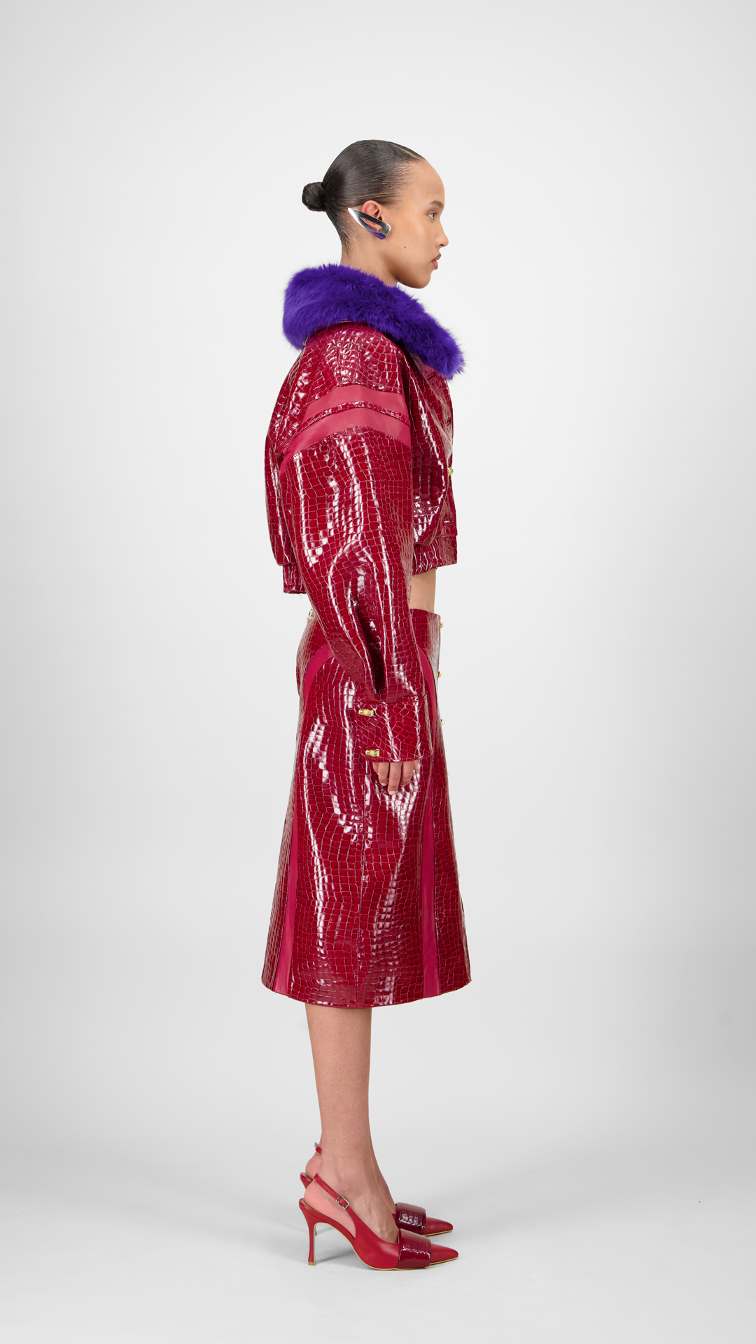 A woman model wearing a red crocodile bomber jacket with a detachable purple faux fur collar and metallic bamboo-like buttons.