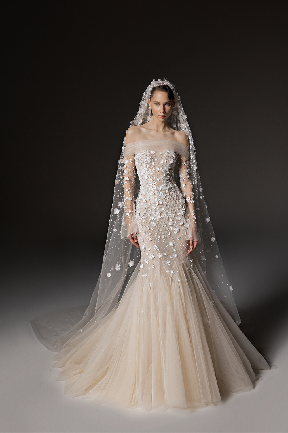 Off-shoulder bridal gown in tulle with sheer long sleeves, three-dimensional floral embroidery and a flared godet skirt.