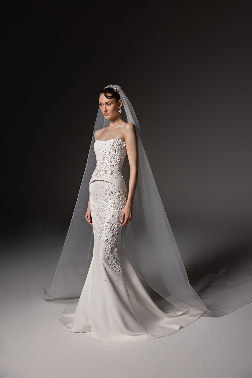 Strapless bridal gown with a sculpted bodice, cascading crystal embellishments, fitted floor-length skirt and sheer tulle veil.