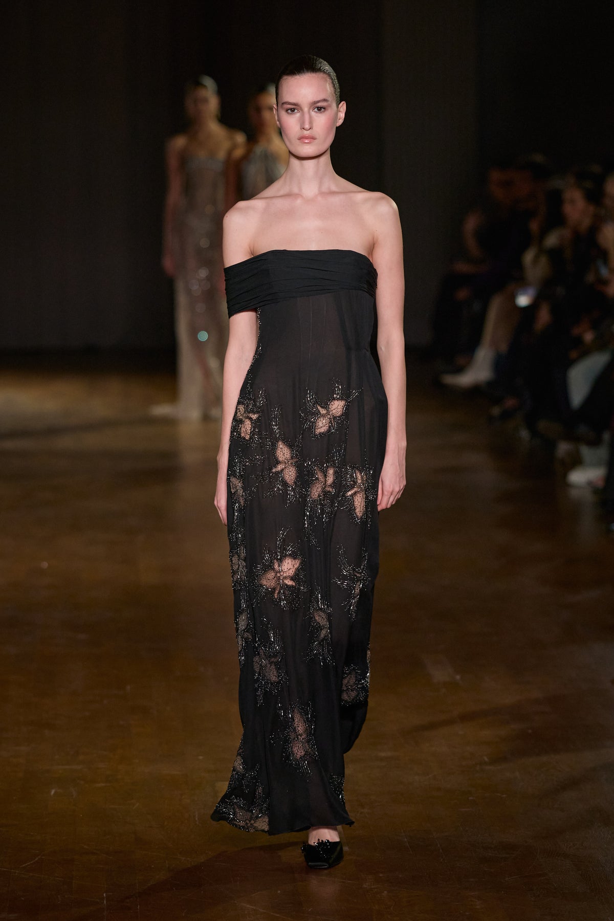 Black floor-length gown with an asymmetric off-shoulder neckline and sheer floral embroidered detailing.