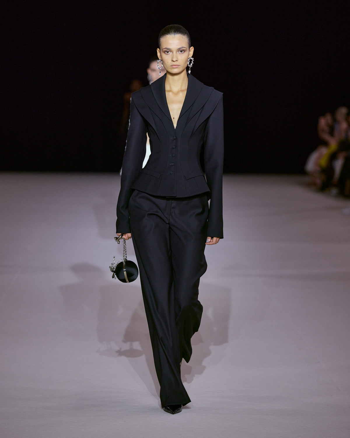 Black tailored jacket with structured shoulders, layered lapels and a sculpted cinched waist, paired with fluid black trousers.