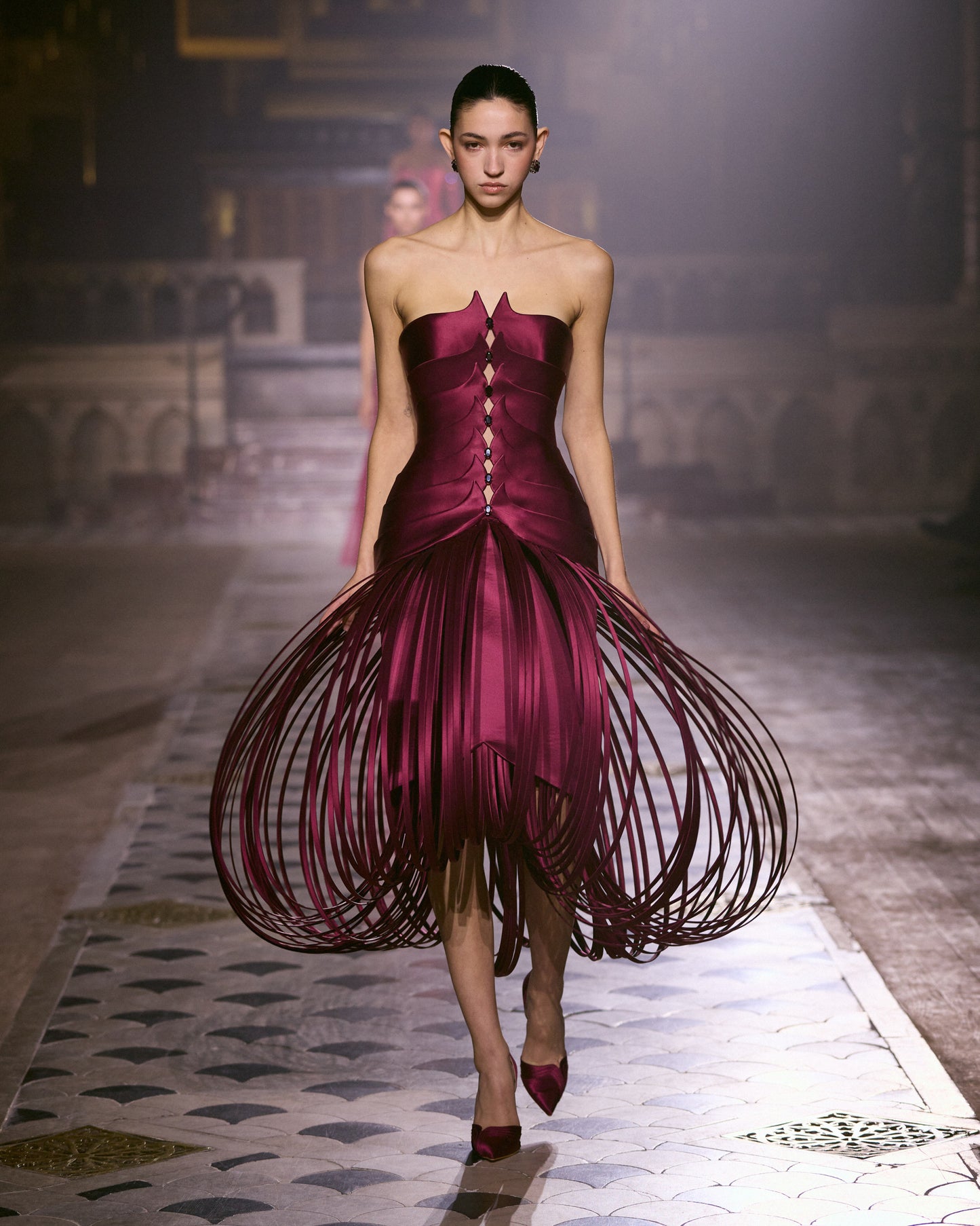 Model walks the runway in Georges Hobeika Spring–Summer 2026 Look 33, wearing a strapless burgundy dress with a flowing, sculptural design.