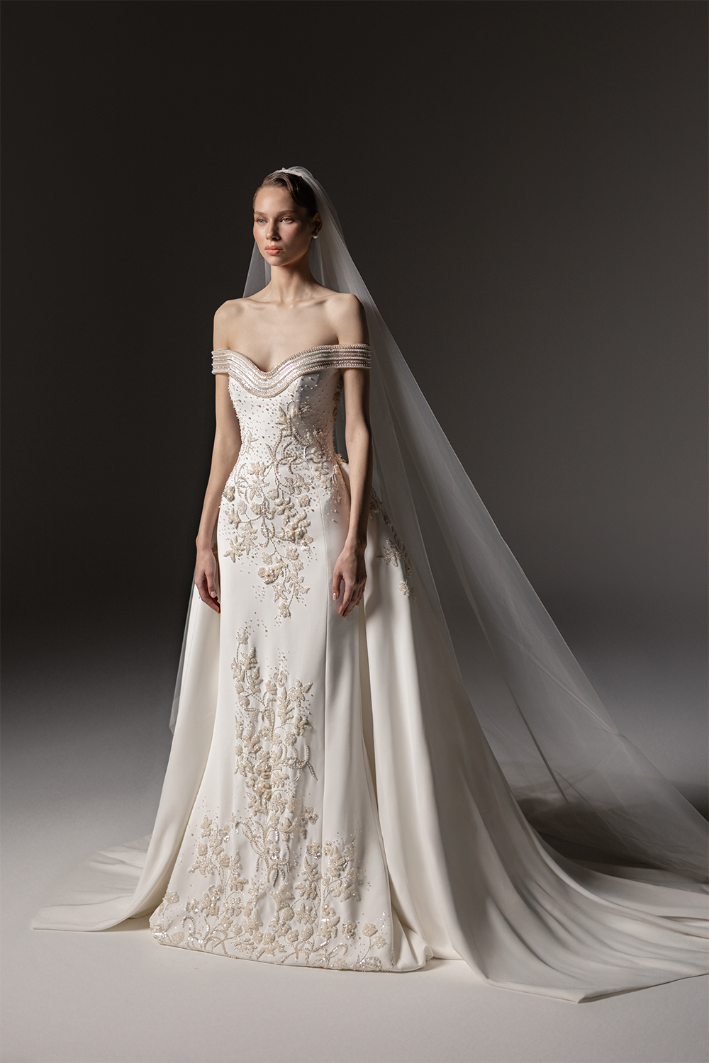 Off-shoulder bridal gown with a sculpted bodice, intricate floral embellishment, floor-length skirt and sheer tulle veil.