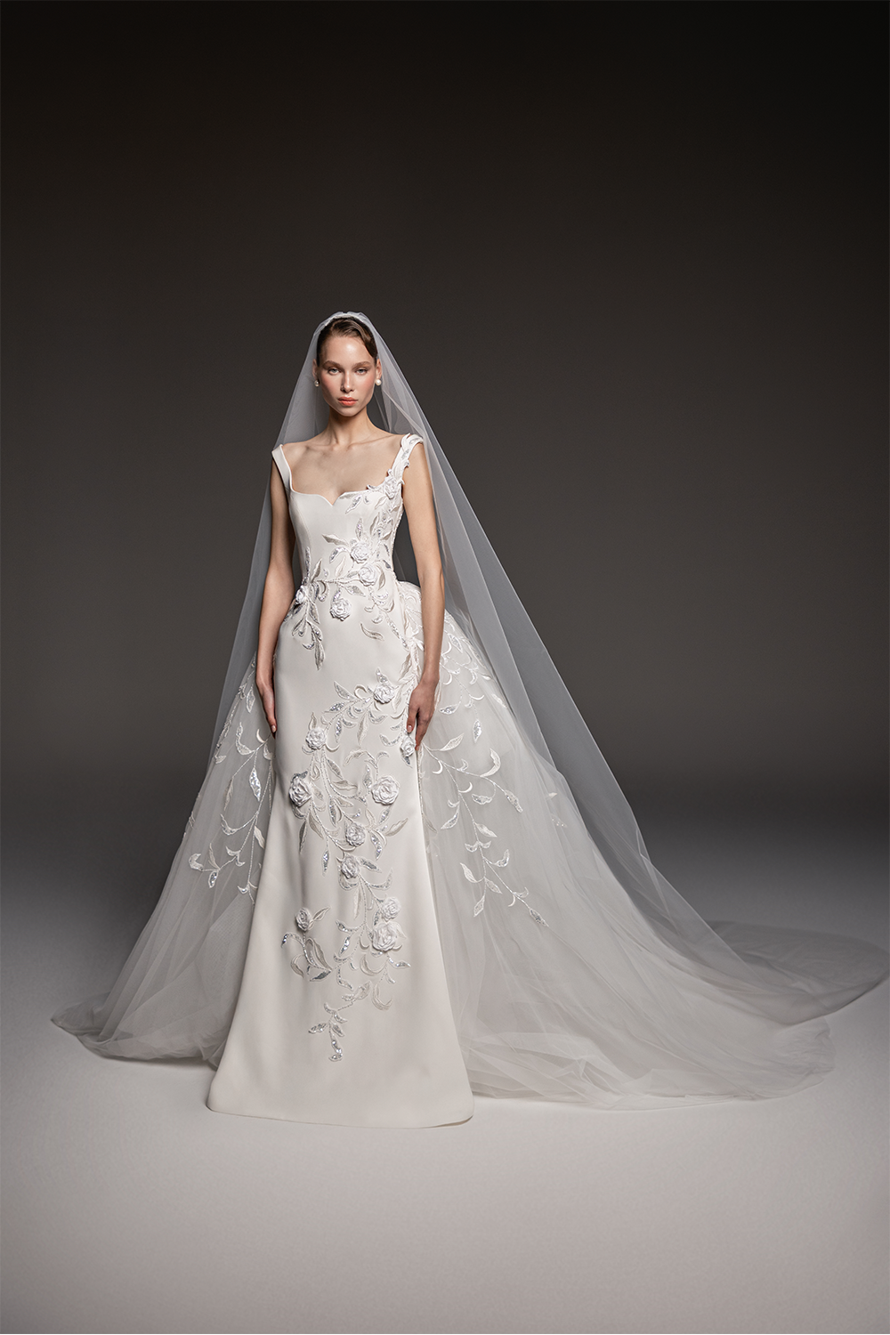 Sculpted bridal gown with floral embroidery, asymmetric shoulder detailing, a fitted silhouette and flowing tulle overskirt.
