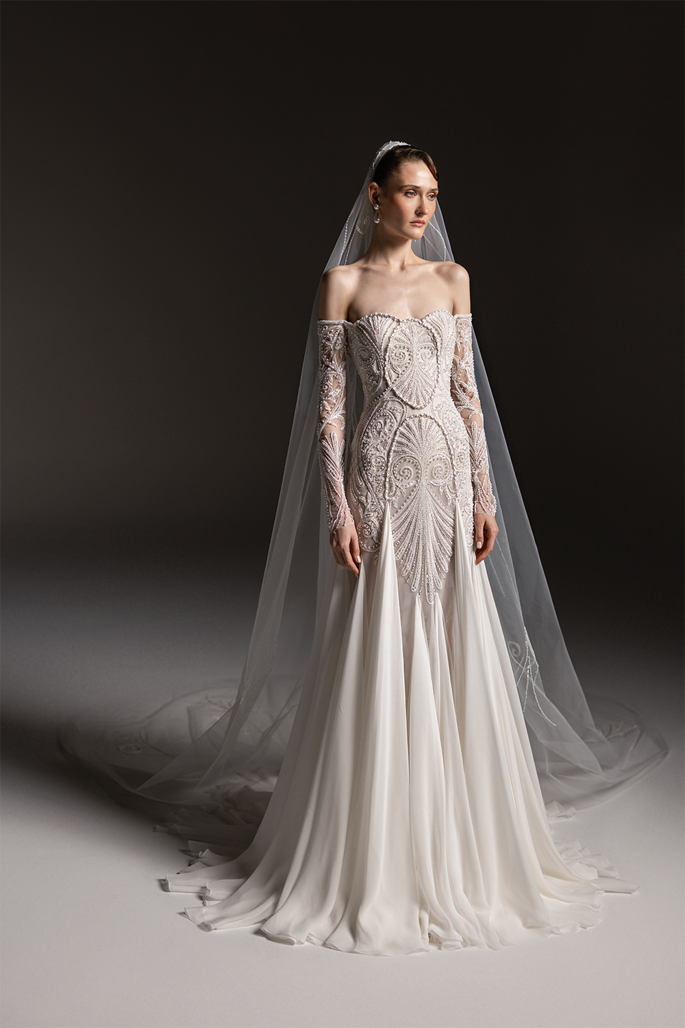 Off-shoulder bridal gown with sheer embroidered sleeves, an intricately beaded bodice and a flowing floor-length skirt.