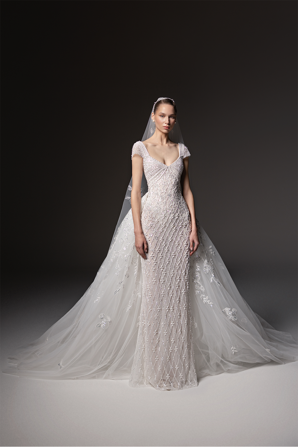 Beaded bridal gown with cap sleeves, a V-neckline, geometric embellishment and a voluminous floral-embroidered tulle overskirt.