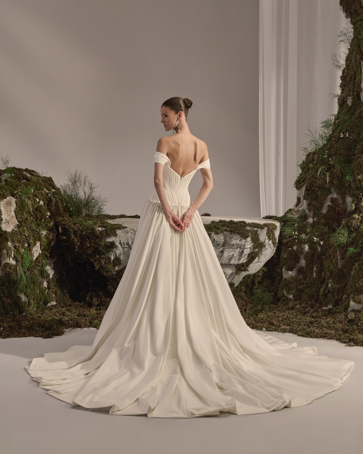 Off-shoulder bridal gown with a sculpted pleated corset bodice, voluminous flowing skirt and sheer veil.