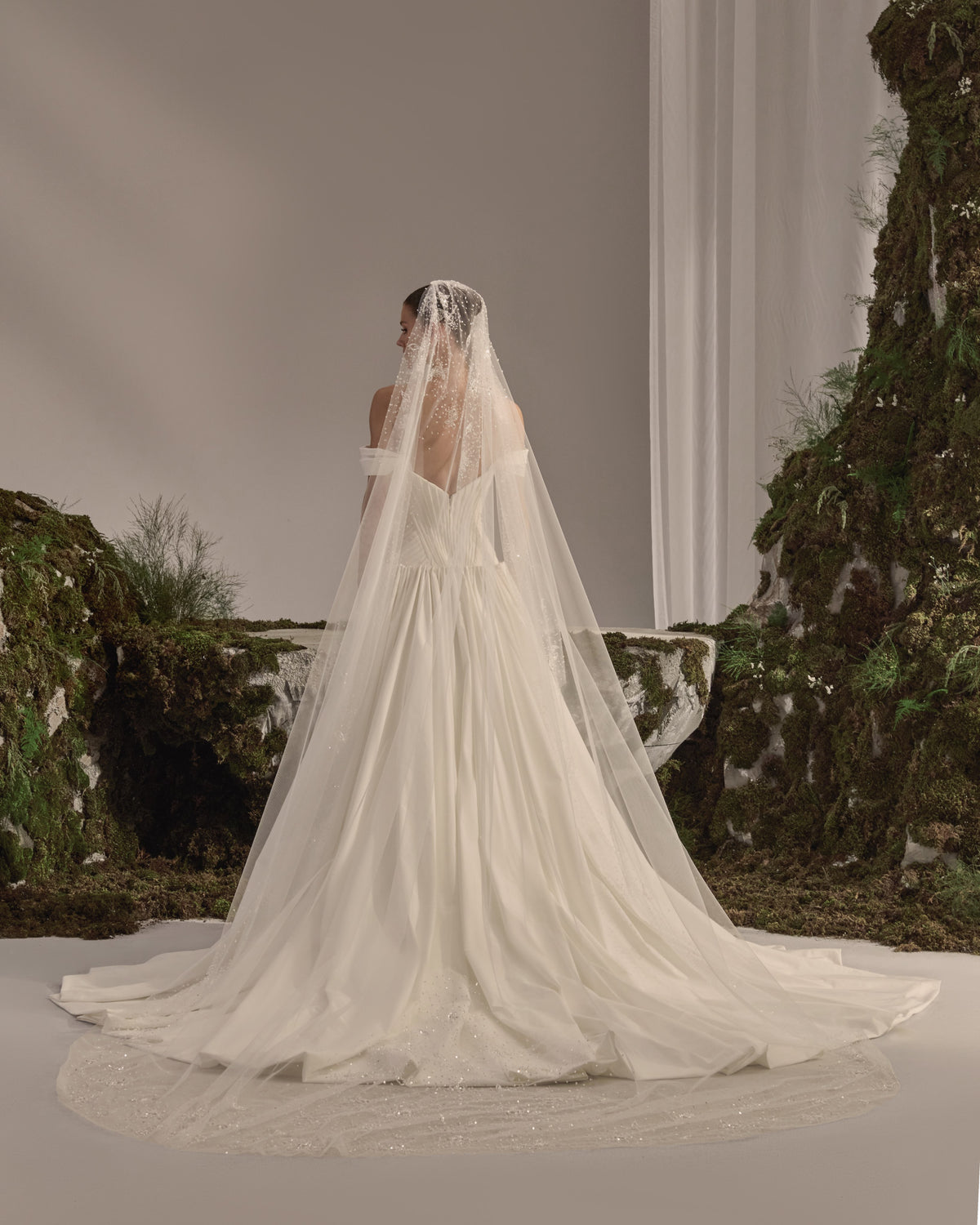 Off-shoulder bridal gown with a sculpted pleated corset bodice, voluminous flowing skirt and sheer veil.