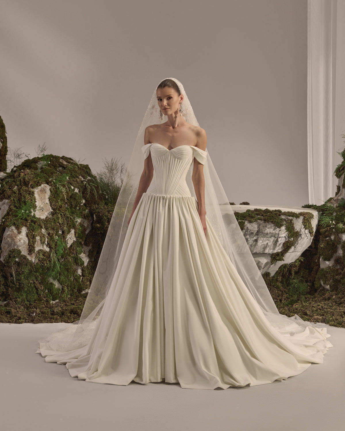 Off-shoulder bridal gown with a sculpted pleated corset bodice, voluminous flowing skirt and sheer veil.