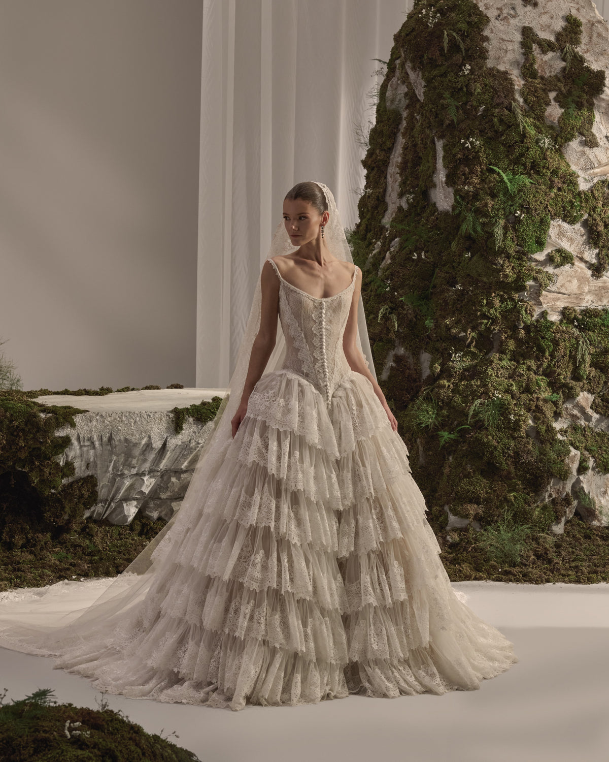 Bridal gown with a fitted lace corset bodice, delicate straps, button detailing, voluminous tiered lace skirt and sheer veil.