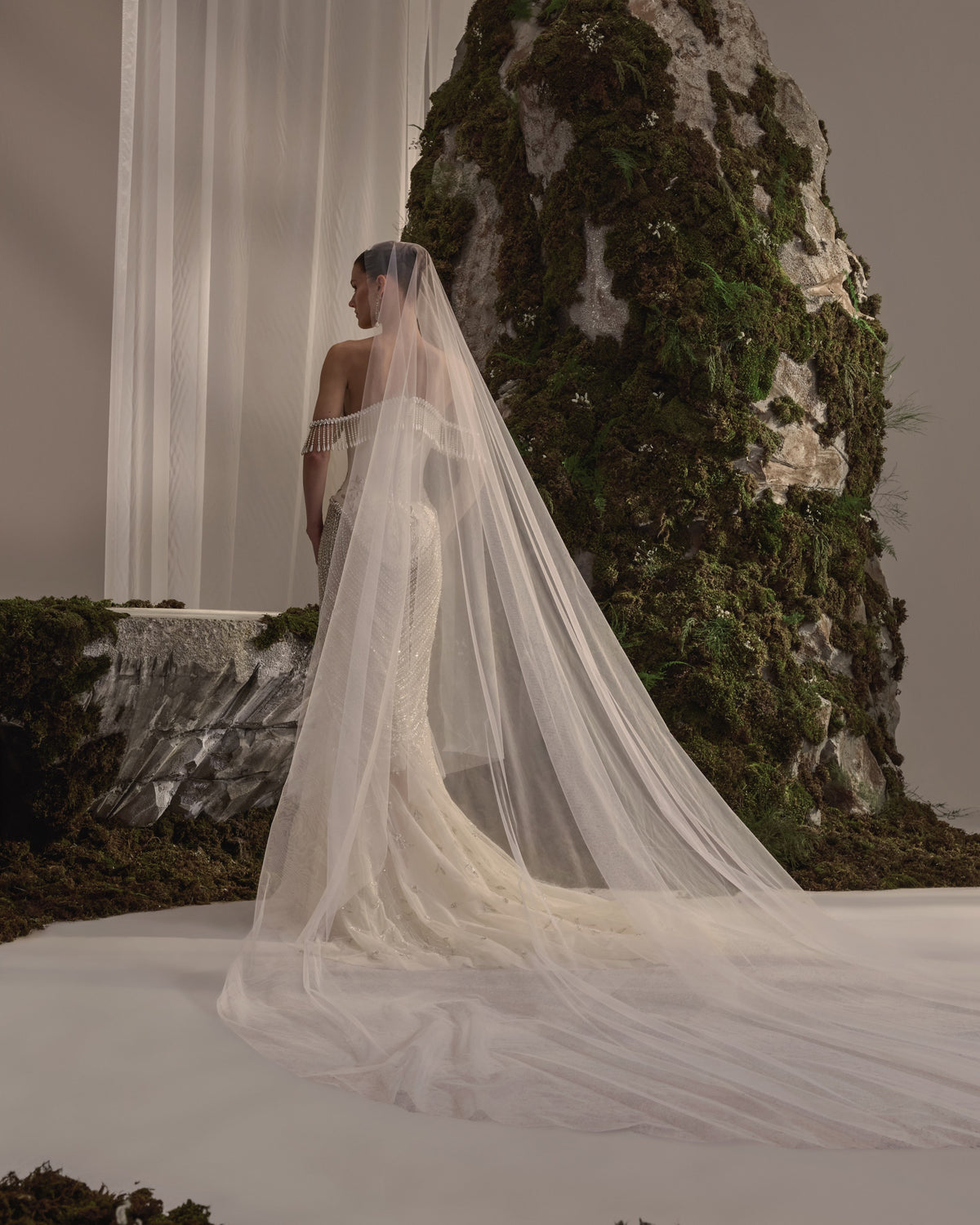 Off-shoulder bridal gown with a sculpted corset bodice, cascading bead embellishment, fitted embroidered skirt and sheer veil.