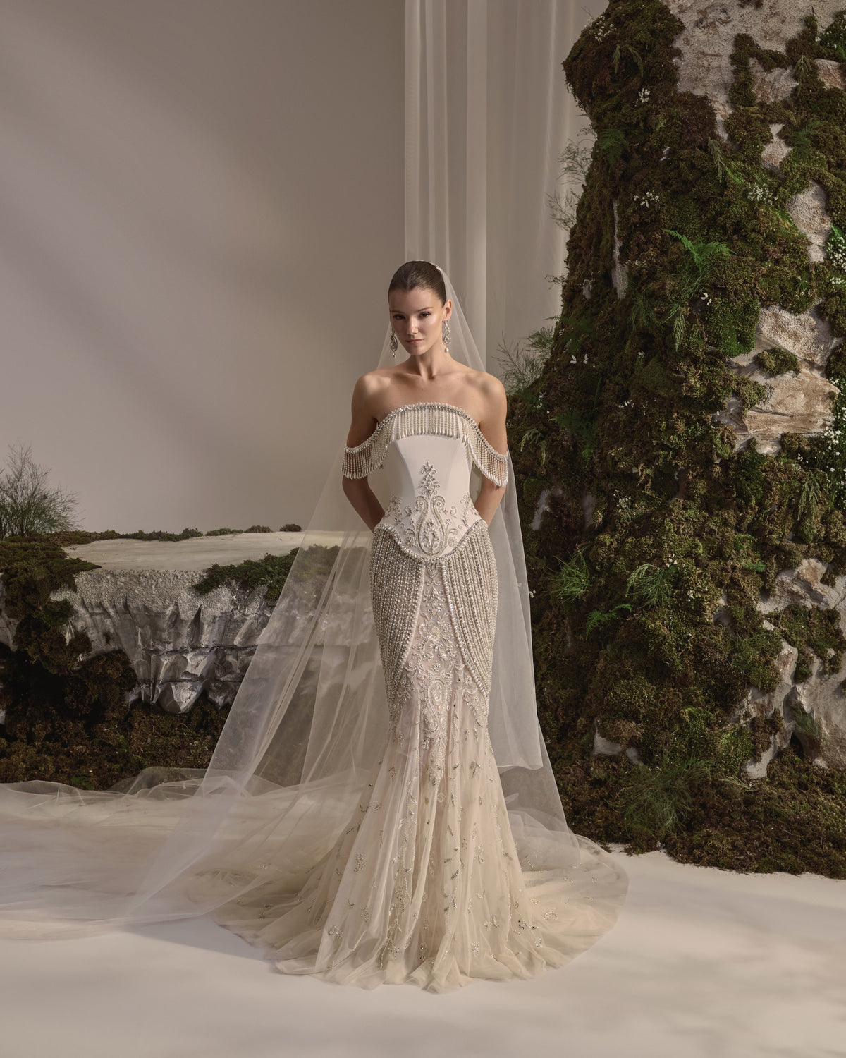 Off-shoulder bridal gown with a sculpted corset bodice, cascading bead embellishment, fitted embroidered skirt and sheer veil.