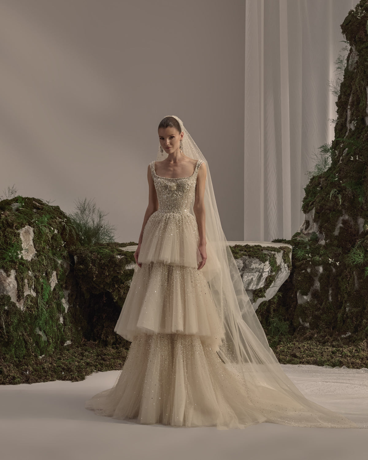 Bridal gown with an embellished square-neck bodice, delicate straps, tiered tulle skirt, shimmering details and a sheer veil.