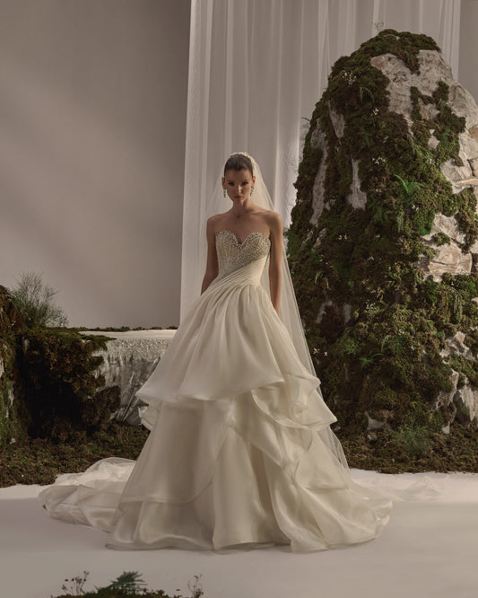 Off-shoulder bridal gown with an embellished draped neckline, fitted silhouette, voluminous lace overskirt and long sheer veil.