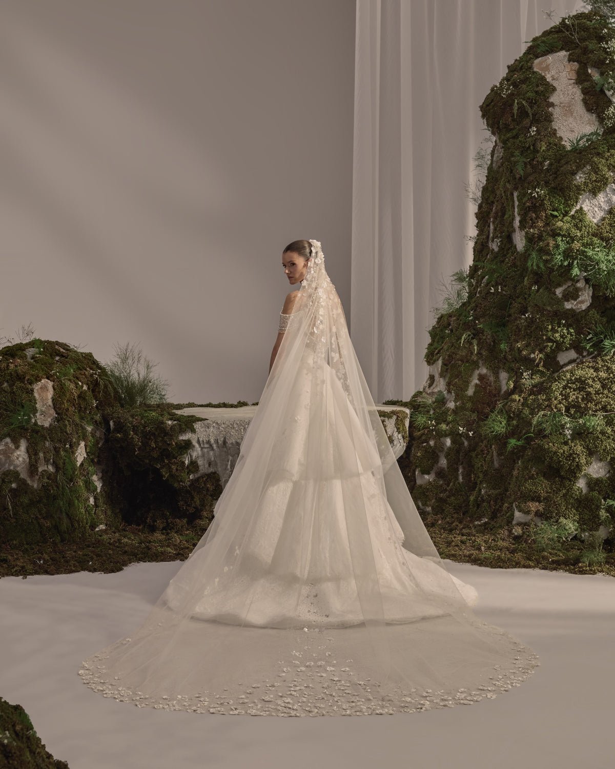 Off-shoulder bridal gown with an embellished draped neckline, fitted silhouette, layered lace overskirt and sheer veil.