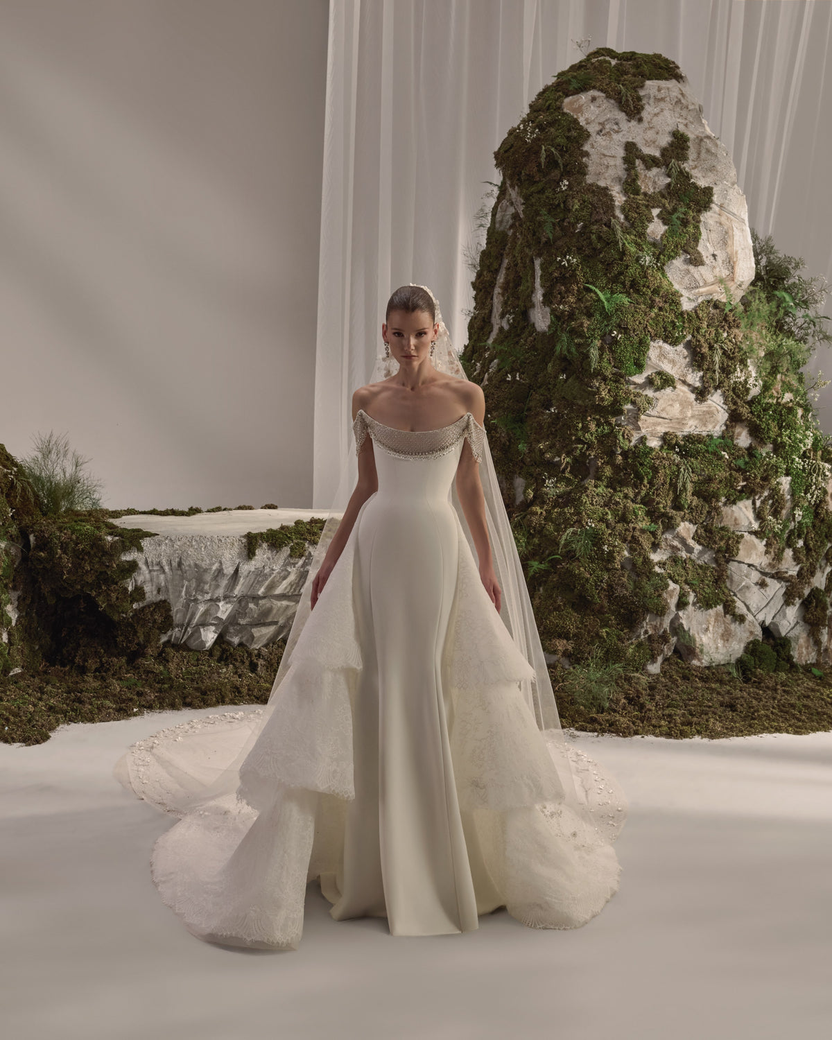Off-shoulder bridal gown with an embellished draped neckline, fitted silhouette, layered lace overskirt and sheer veil.