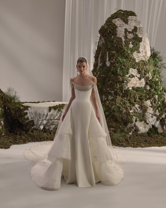 Off-shoulder bridal gown with an embellished draped neckline, fitted silhouette, layered lace overskirt and sheer veil.