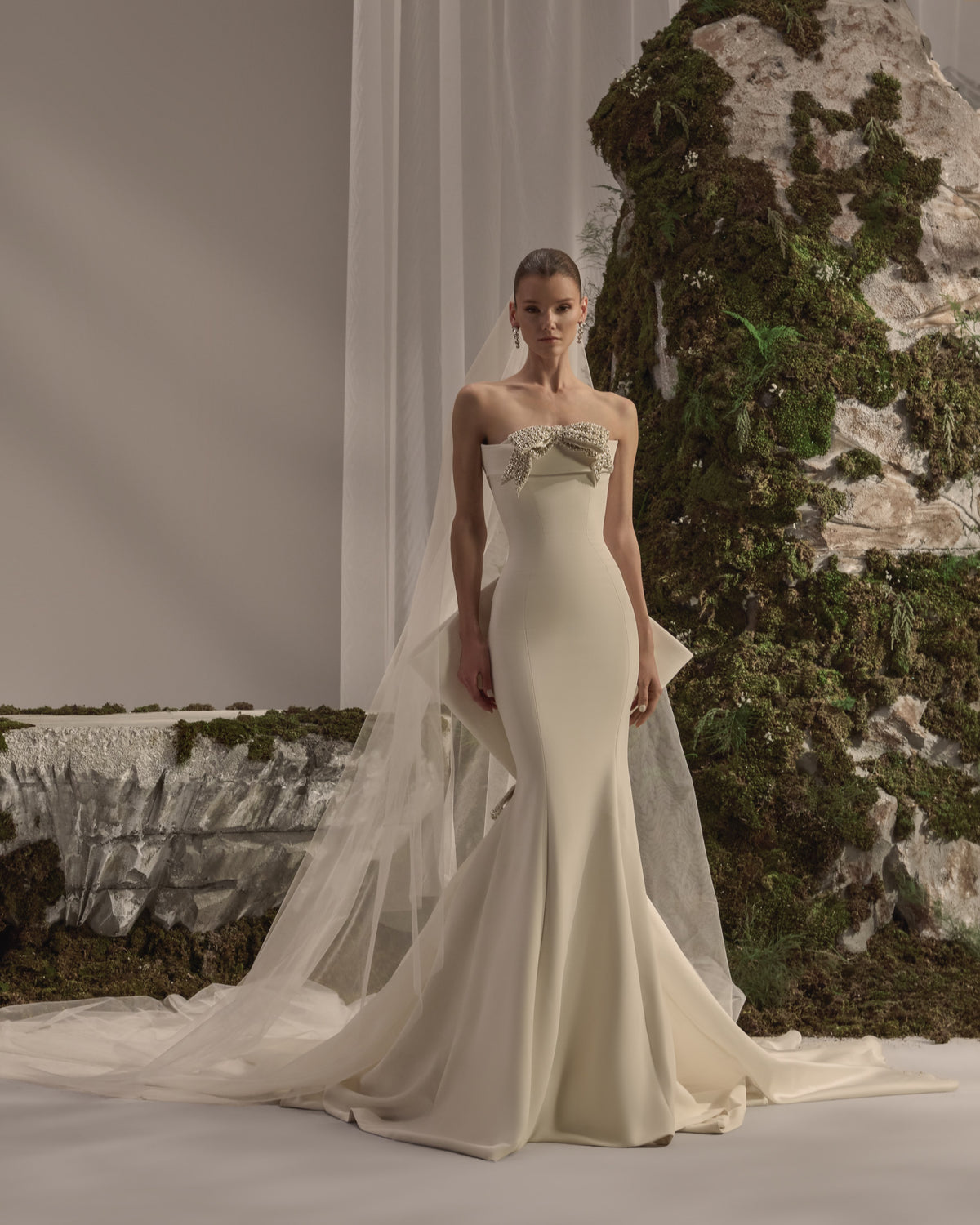 Strapless bridal gown with an embellished bow neckline, fitted sculpted silhouette and flowing train with a sheer veil.