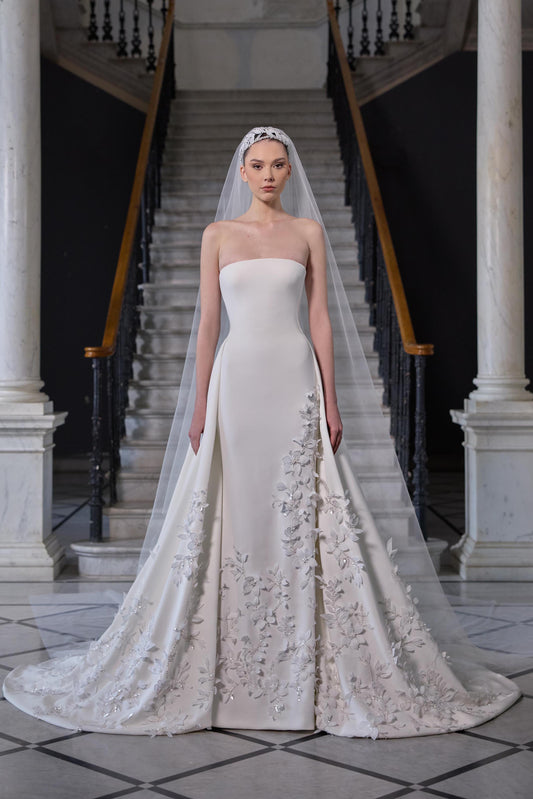 Strapless bridal gown with a sculpted bodice, voluminous floor-length skirt, delicate floral embroidery and sheer tulle veil.