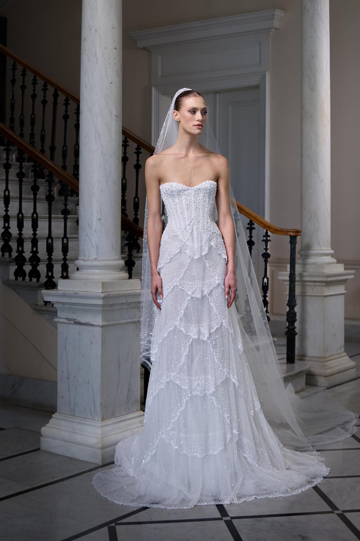 Strapless bridal gown with a beaded corset bodice, layered scalloped tulle skirt and sheer embellished veil.