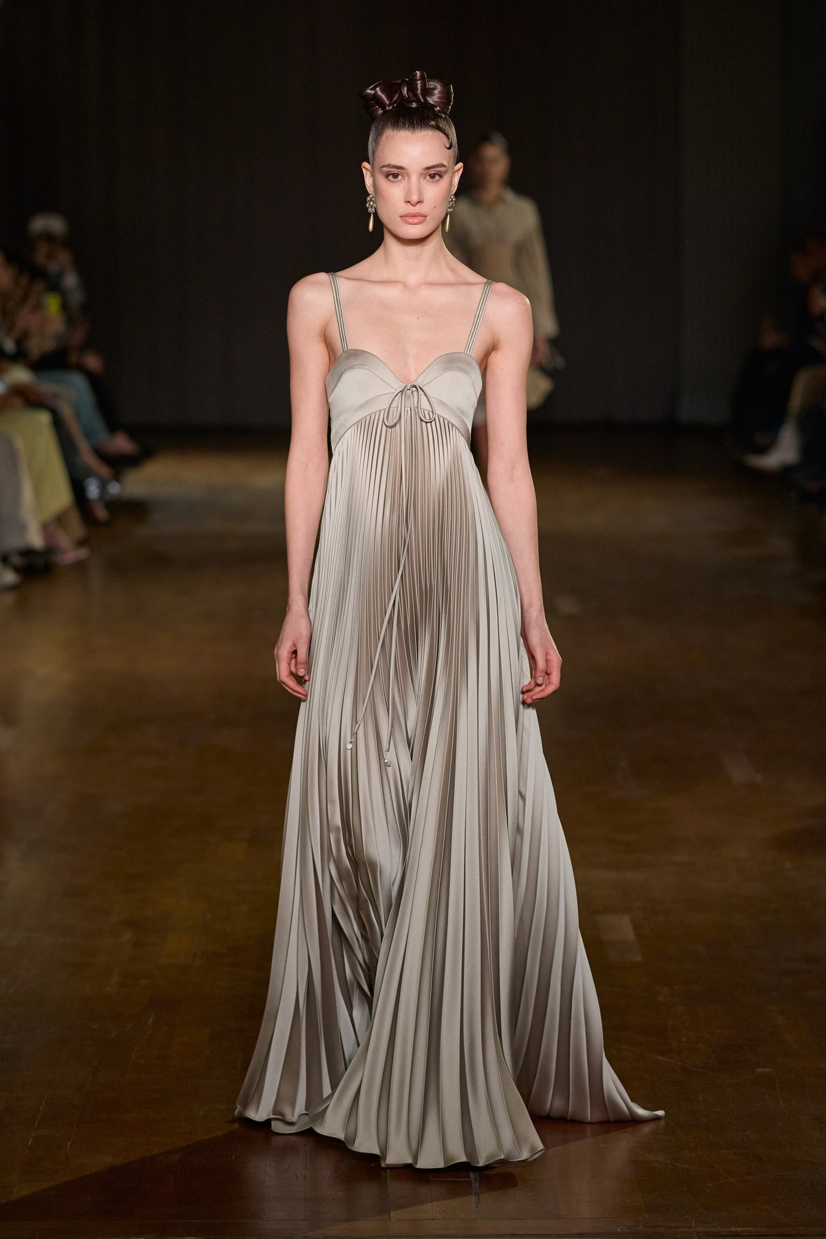 Floor-length pleated satin gown with structured bust, thin double straps, and delicate tie detailing at the neckline.