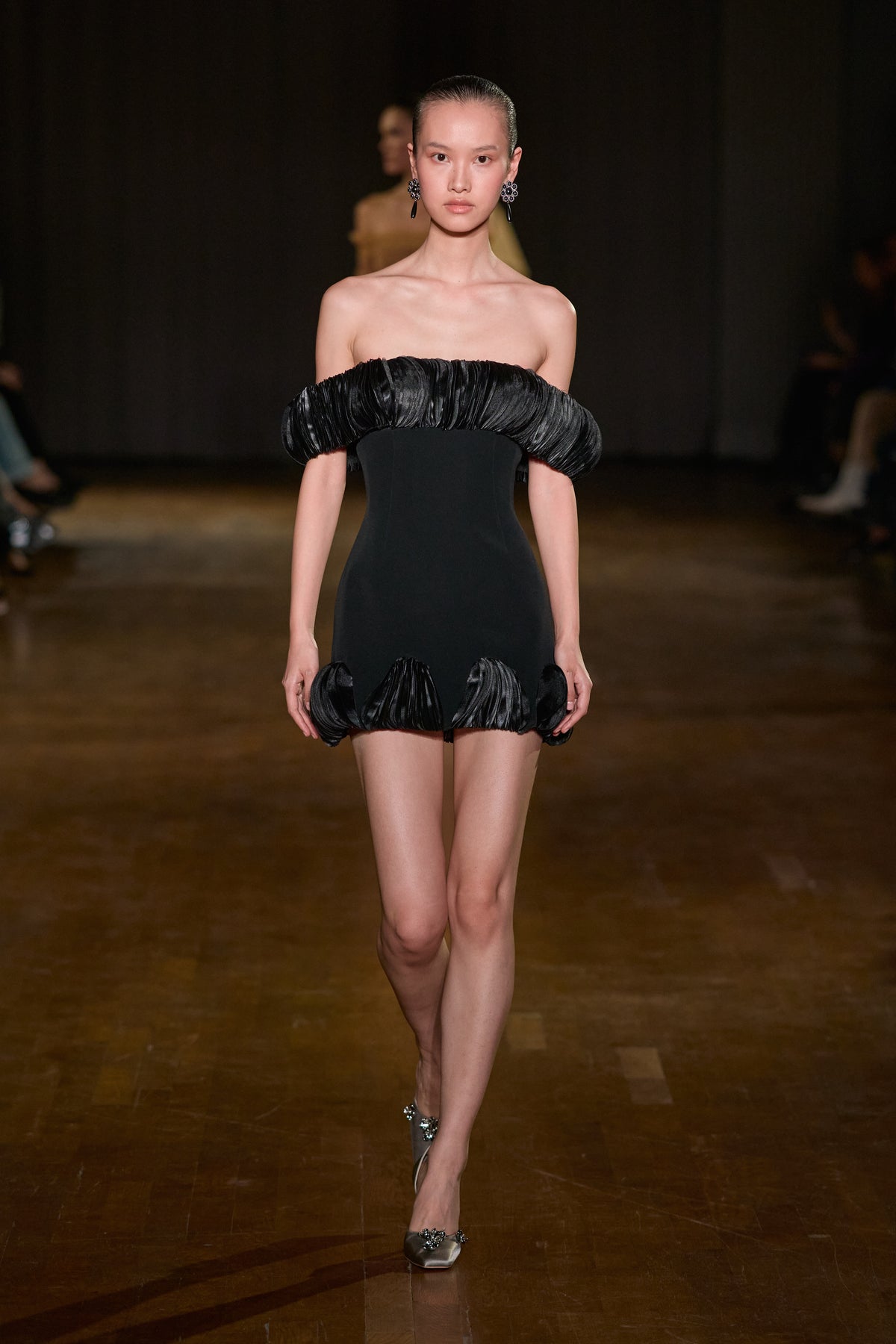 Black fitted mini dress with a sculptural pleated off-shoulder neckline and matching pleated detailing along the hem.