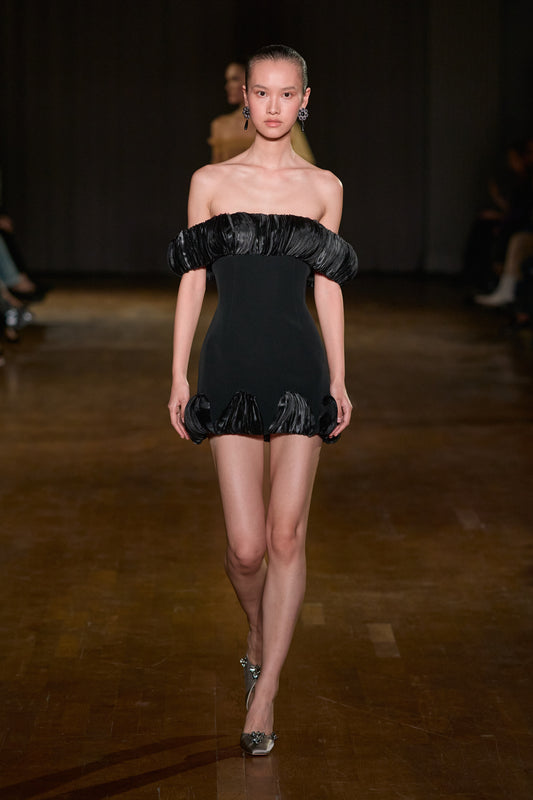 Black fitted mini dress with a sculptural pleated off-shoulder neckline and matching pleated detailing along the hem.
