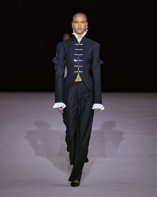Black tailored jacket with sculpted sleeves, crystal embellishments and white ruffled cuffs, paired with fluid black trousers.